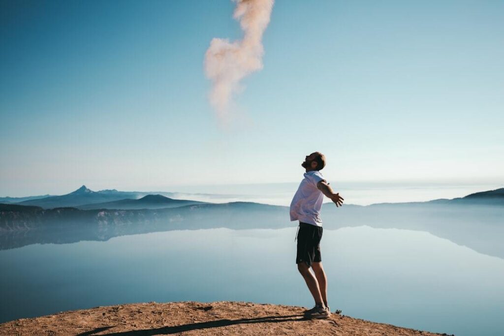Man standing on mountain cliff with arms outstretched, facing sky and lake below