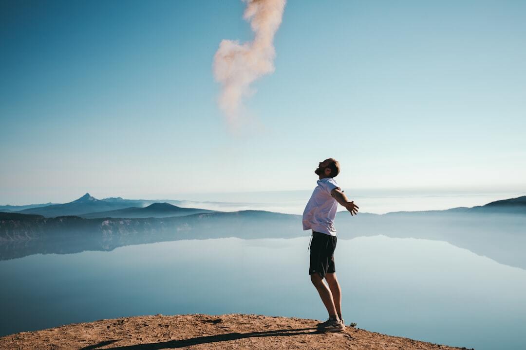 Man standing on mountain cliff with arms outstretched, facing sky and lake below