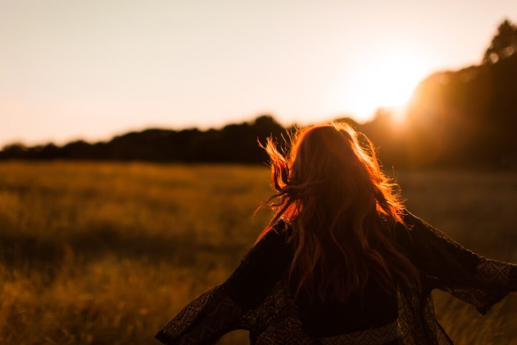 Woman with red hair walking through a golden field at sunset with arms open wide